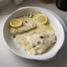 Baked cod filet served with a rich lemon cream sauce on a white plate.