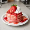 Fluffy strawberry pancakes topped with fresh strawberries and syrup on a plate.